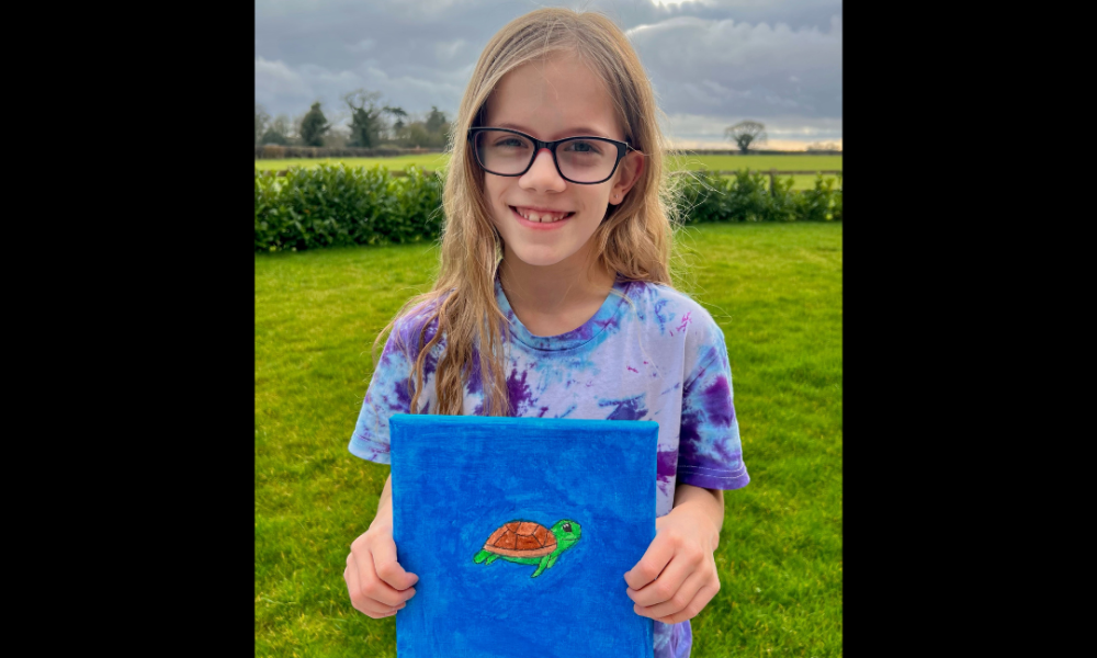 Young girl holding a blue painting with a green turtle, stood in a green field