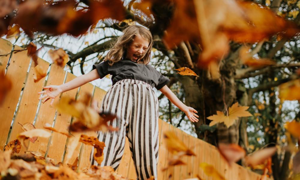 Autumn leaves scatter, falling towards the camera, surrounding a girl as she laughs after throwing a large handful into the air. Depicts a carefree childhood, and autumn time. 