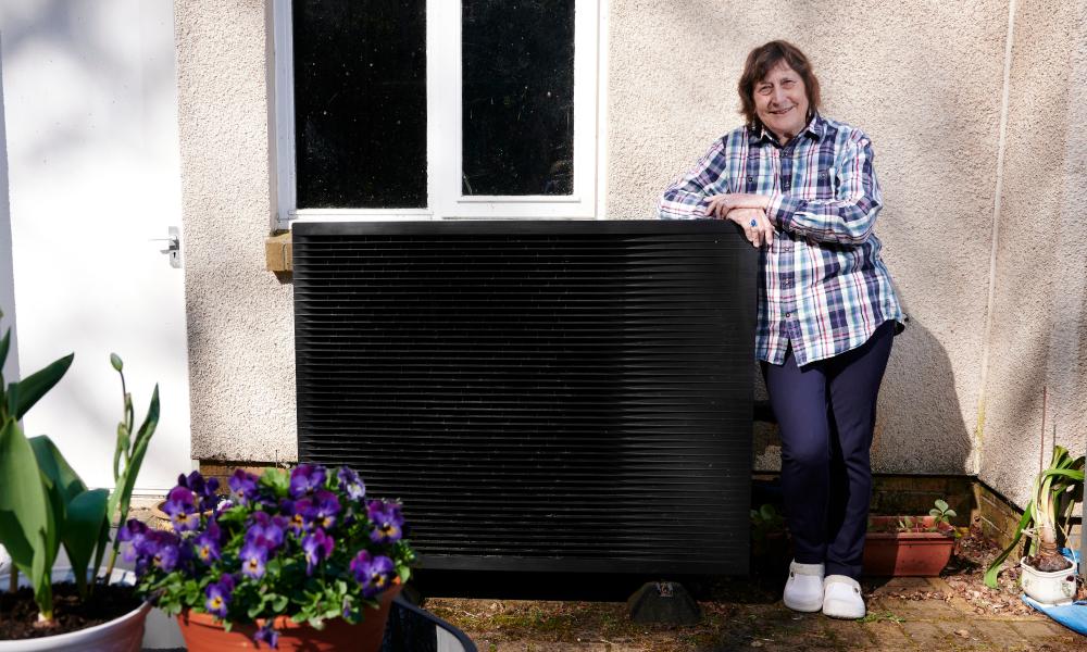 Lesley stood proudly with her heat pump. The heat pump is beside the outside of her home and surrounded by plant pots with bright purple flowers.