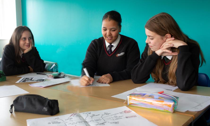 Three school pupils sat at around a table writing on paper